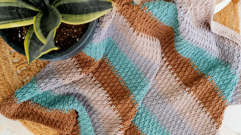 Alpine Stitch Blanket in stripes of tan, light blue and gray, draped over a woven basket beside a snake plant