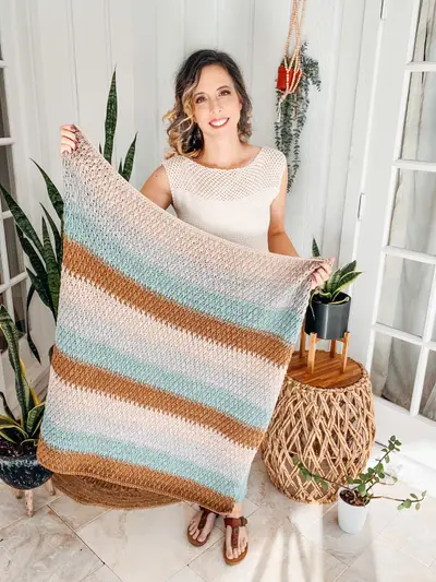 Woman holding up the finished Alpine Stitch Blanket in tan, gray and blue stripes