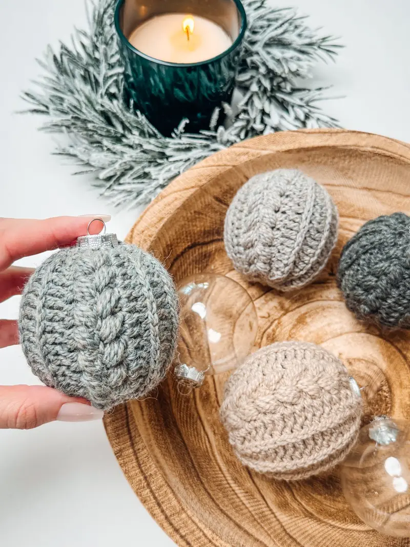 Hand holding a gray Autumn Wheat Bauble Ornament over a wooden bowl of crochet ornaments