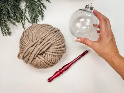 Hand holding a clear ornament shell beside a skein of oatmeal yarn and a crochet hook for the Autumn Wheat Bauble