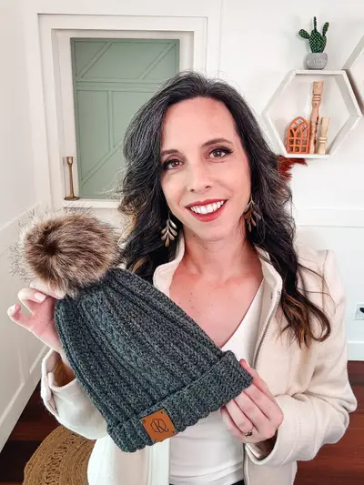 Woman holding a charcoal Autumn Wheat Flat Crochet Hat with a faux fur pom pom