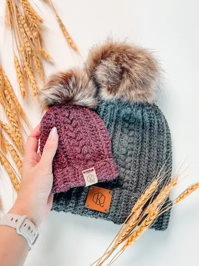 Hand holding plum and teal Autumn Wheat Flat Crochet Hats with pom poms