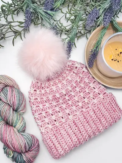 Pink Block Stitch Beanie with a large pink pom pom beside a speckled skein of yarn