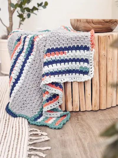 Block Stitch Crochet Blanket in gray, navy, coral and teal stripes folded on a wooden stool