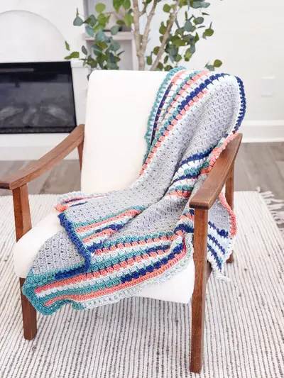 Striped Block Stitch Crochet Blanket in gray, navy, coral and teal draped over a white chair
