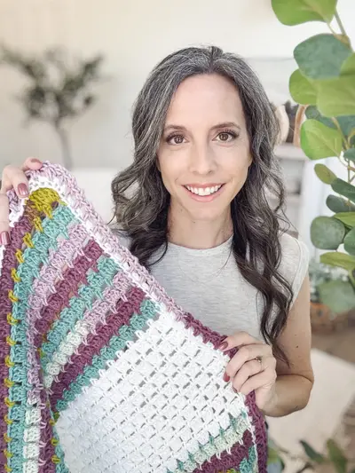 Woman holding up the Block Stitch Crochet Blanket in white with purple, teal and mustard stripes