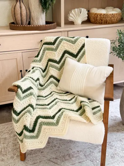 Chevron Petal Loop Blanket in cream and green zigzag stripes on an armchair with a pillow