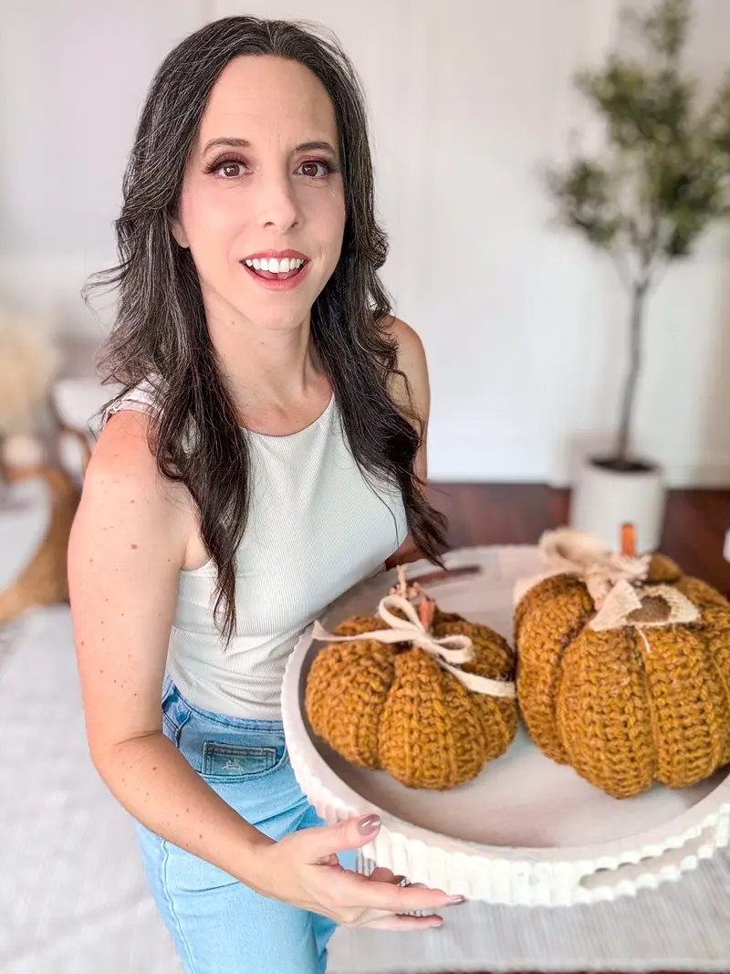 Smiling woman holding a white tray of two mustard crochet pumpkins tied with ribbon bows