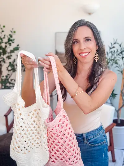 Smiling woman holding up two Circle Leaf Crochet Market Bags in cream and pink