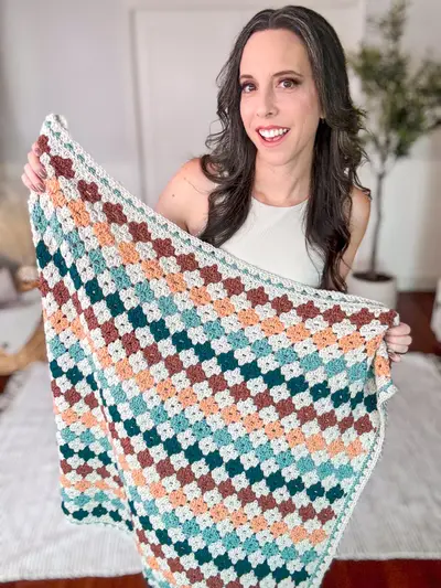 Woman holding up the Colorful Hues Crochet Blanket in teal, peach, rust and cream granny stitch