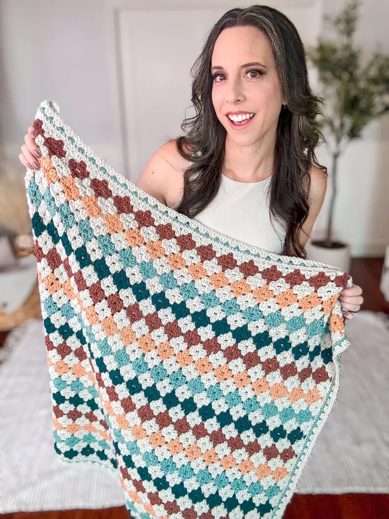 Woman holding up the Colorful Hues Crochet Blanket in teal, peach, rust and cream granny stitch