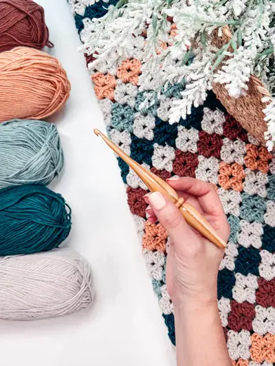 Hand crocheting the Colorful Hues Crochet Blanket beside skeins of peach, teal and gray yarn