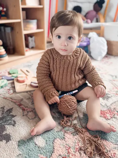 Toddler sitting on a rug wearing a brown Easy Cuff to Cuff Child Crochet Sweater