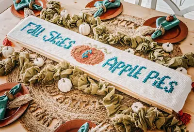 Cream table runner reading Fall Skies Apple Pies in teal around an orange pumpkin
