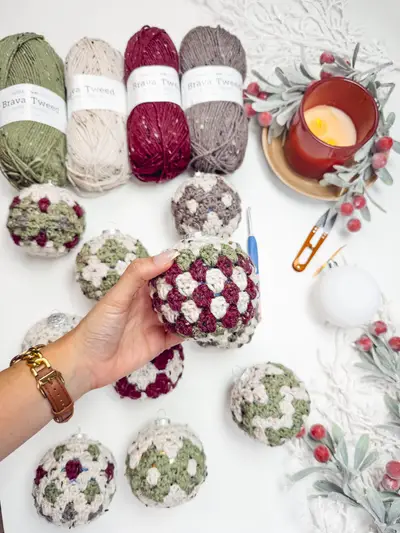 Hand holding a Granny All the Way Ornament in burgundy, sage and cream with yarn skeins