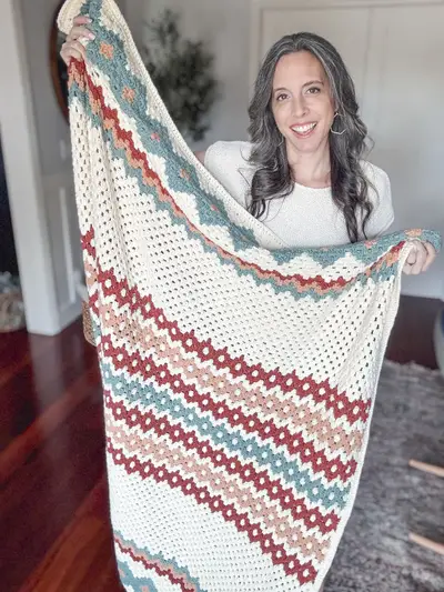 Woman holding up the cream Gemstone Hues Crochet Blanket with rust, blush and blue stripes