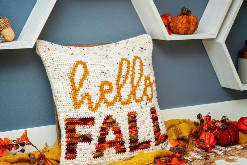Hello Fall Crochet Pillow Cover with gold script and multicolor letters by a blue wall