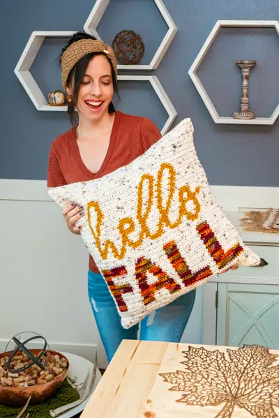 Smiling woman in a rust top holding up the Hello Fall Crochet Pillow Cover