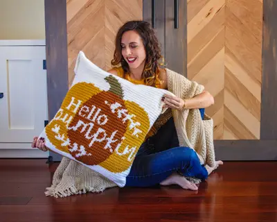 Smiling woman sitting on the floor holding the Hello Pumpkin Crochet Pillow
