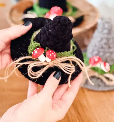 Hand holding a black crochet witch hat with red mushrooms and a twine bow
