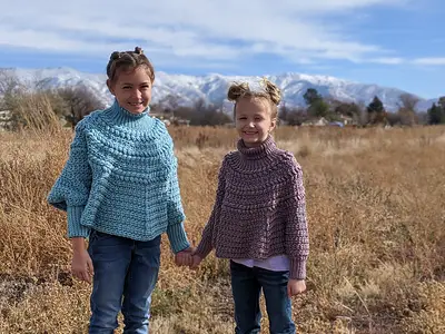 Two girls in blue and mauve Macchiato Crochet Ponchos in a field by mountains