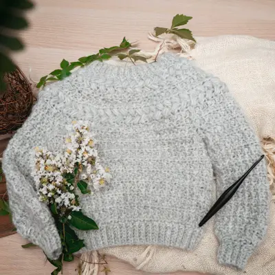 Light gray child Macchiato Crochet Sweater laid flat with a crochet hook and flowers