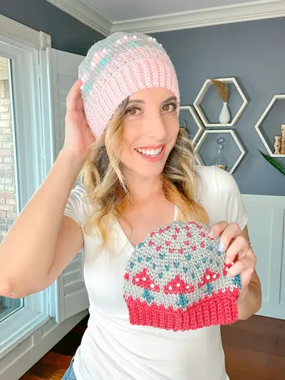 Woman in a pink Mushroom Crochet Beanie holding a red one with mushroom colorwork
