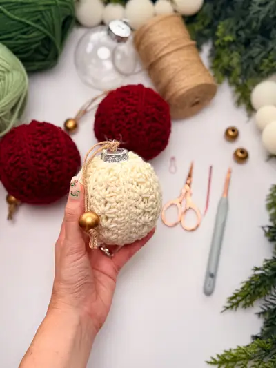 Hand holding a cream Petal Loop Stitch Ornament beside red ones and supplies