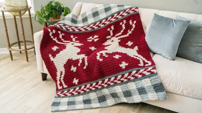 Prancing All the Way Deer Crochet Blanket in red with a cream deer and a gingham border, on a sofa