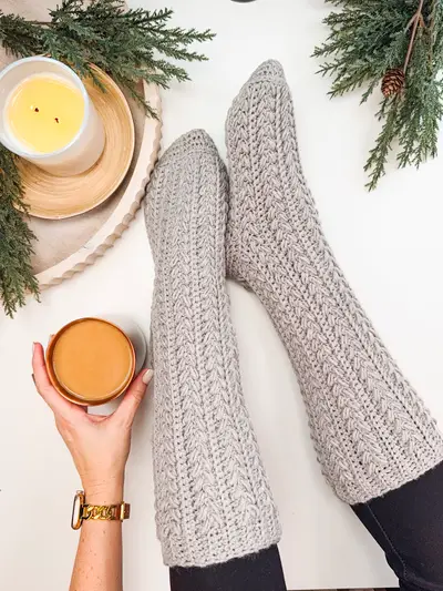 Feet in gray Snowdrift Wheat Slipper Socks holding a cup of tea