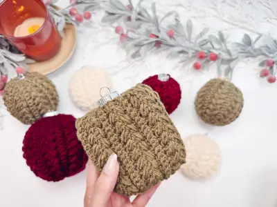 Hand holding an olive Snowy Wheat Ornament with burgundy and cream ones nearby