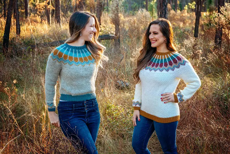 Two women wearing the Traveler Fair Isle Crochet Sweater, one in gray with a blue and gold yoke and one in white with a gold, teal and red yoke