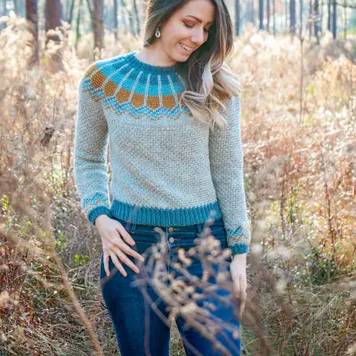 Woman in the gray Traveler Fair Isle Crochet Sweater with teal ribbing, walking through tall grass