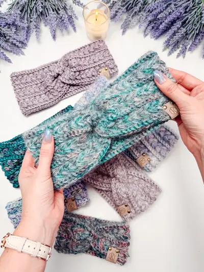 Hands holding a teal Wheat Stitch Ear Warmer beside lavender and gray ones