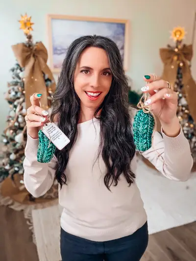 Smiling woman holding up Wheat Stitch Pickle Ornaments by a Christmas tree