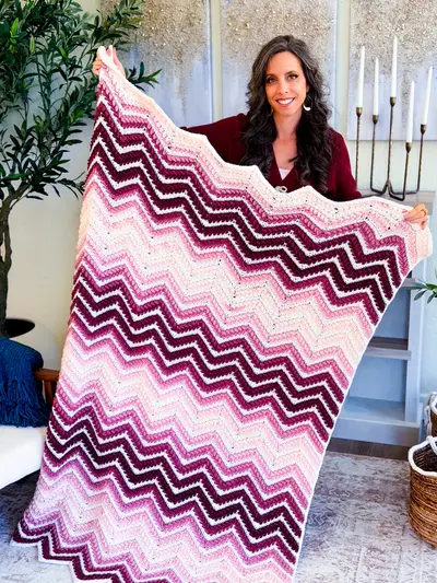 Woman holding up the pink and burgundy Wheat Stitch Ripple Blanket in front of a fireplace